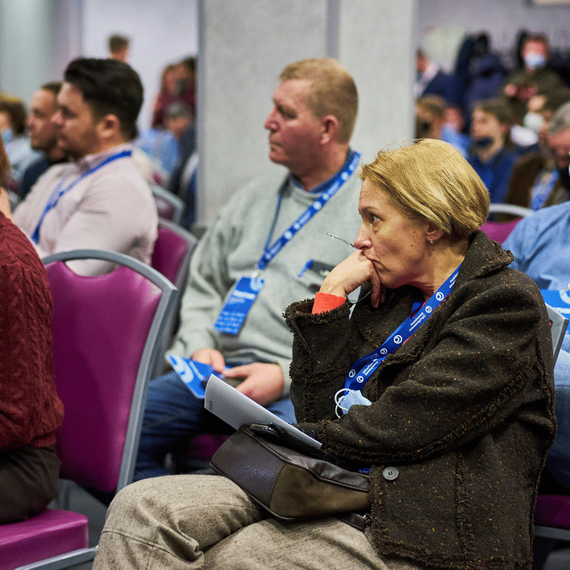 Numerous people sitting inside an event space and listening to a speaker.