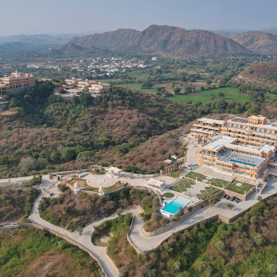 Aerial view of Fateh Vilas, the best resort in Udaipur, surrounded by green mountains with a swimming pool, a road, walking pathways, and lawns