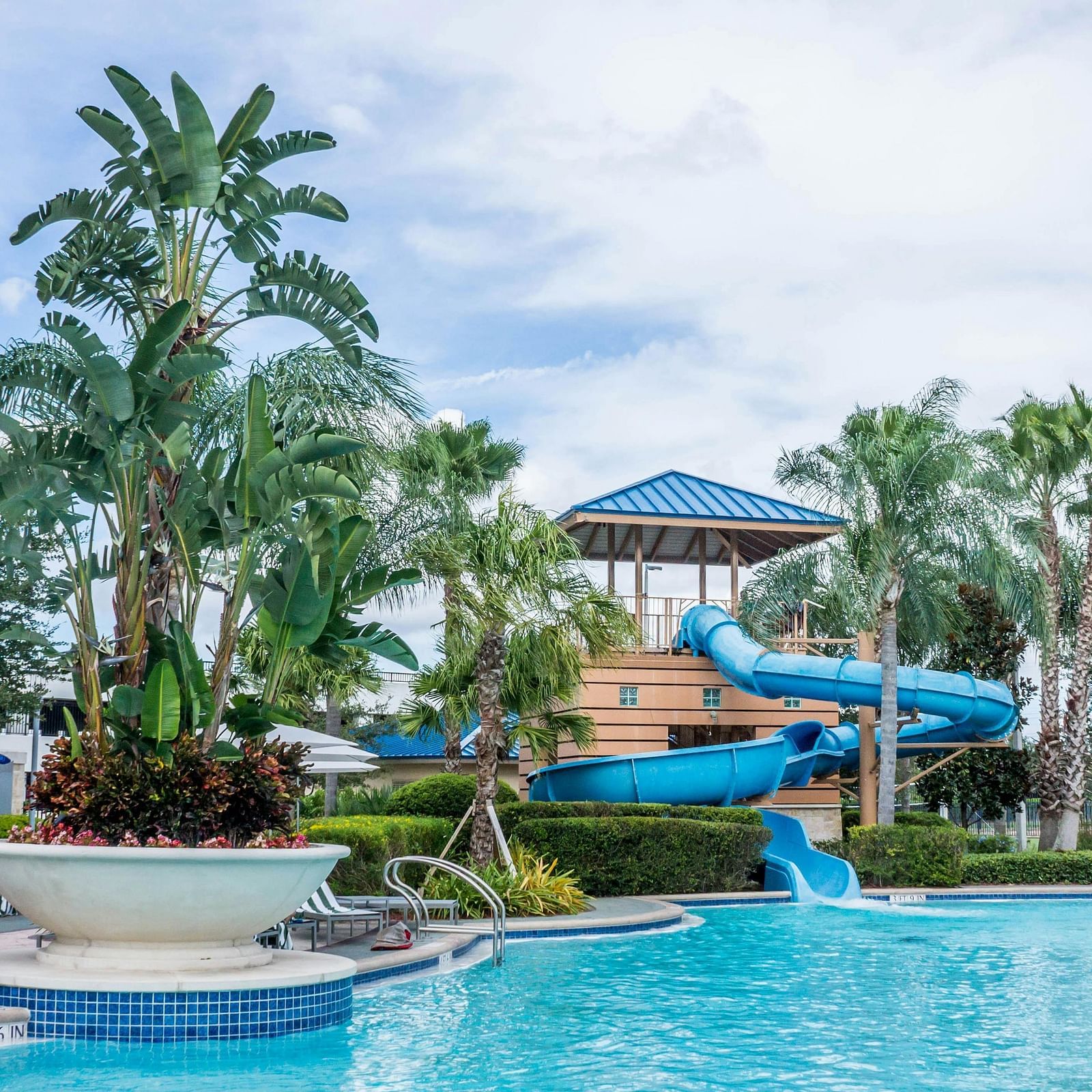 A large, vibrant swimming pool with a waterslide, surrounded by palm trees and lounge chairs at a sunny resort.