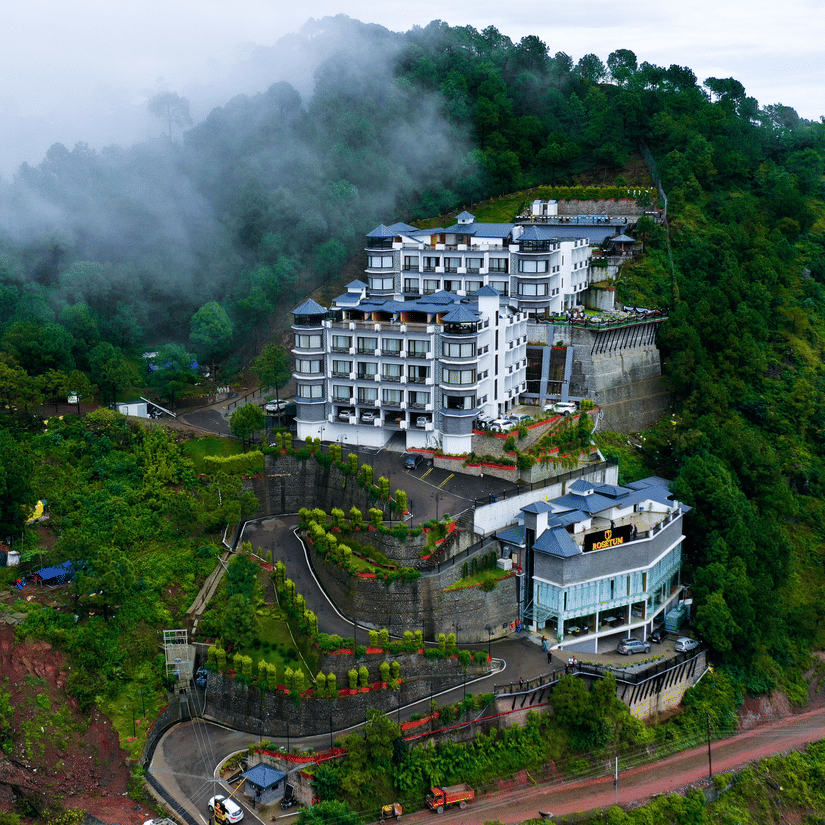 Aerial drone view of Rosetum Hotels Kasauli nestled among misty green hills with terraced landscaping and winding access road