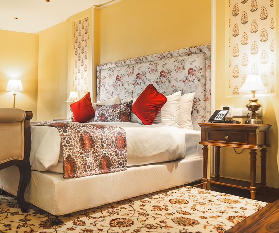 The energised look of the Super deluxe cottage at The Garden Ananta Elite with enchanting white table lamps on both sides, red pillows to up the comfort, and a cushioned seat for two, placed adjacent to the bed.