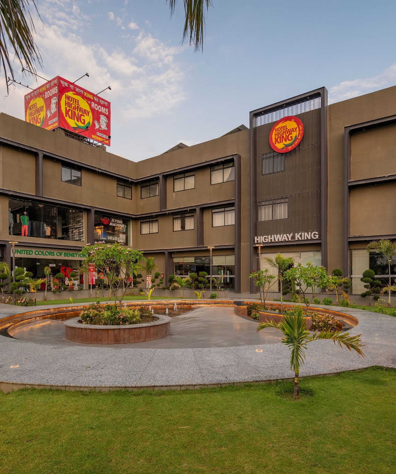 A modern, multi-storey building with a distinctive facade is surrounded by a landscaped garden with a circular fountain in the foreground.