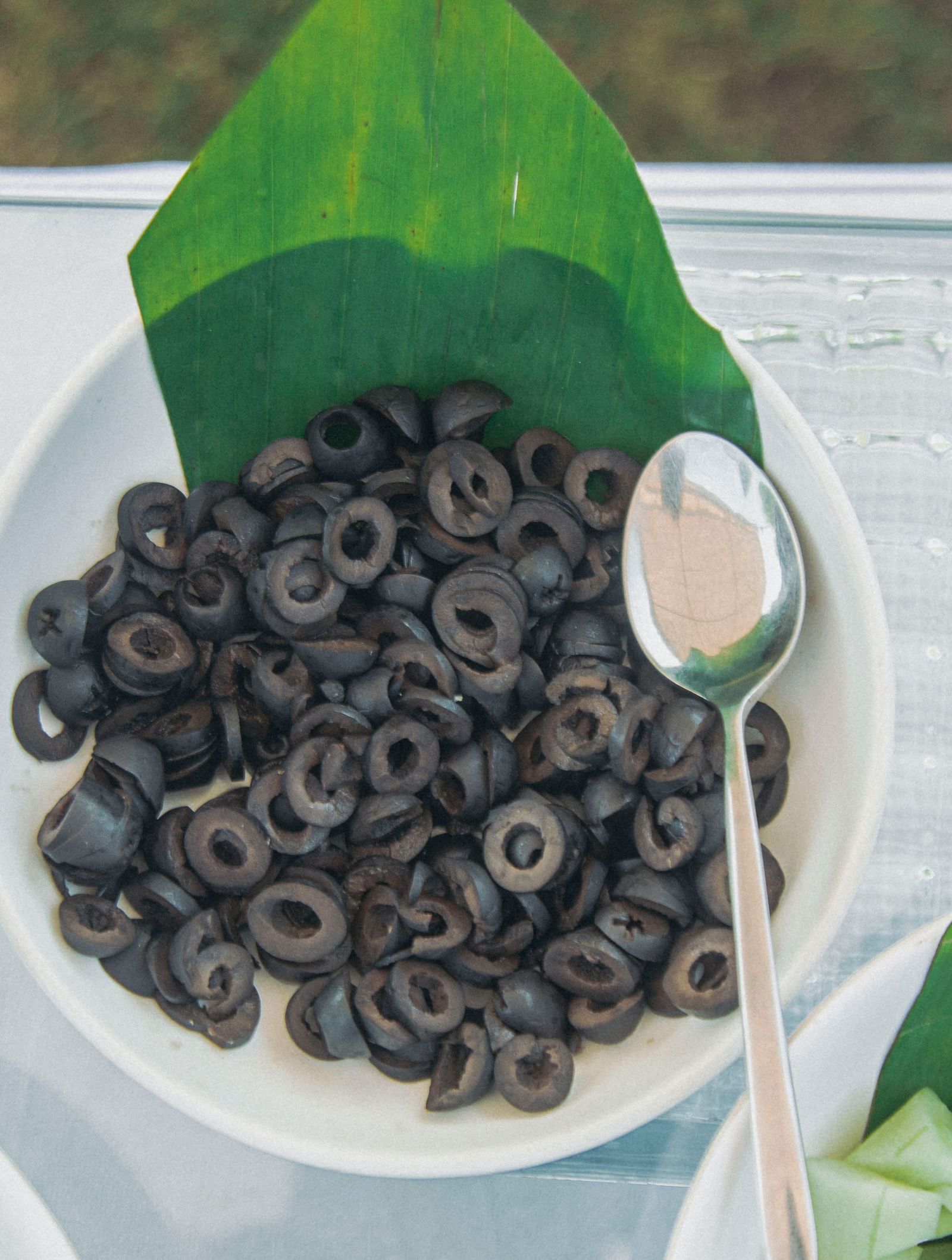 Bowl of black olives placed on a glass counter with a serving spoon and green leaf at Fireflies Resorts, Kabini.