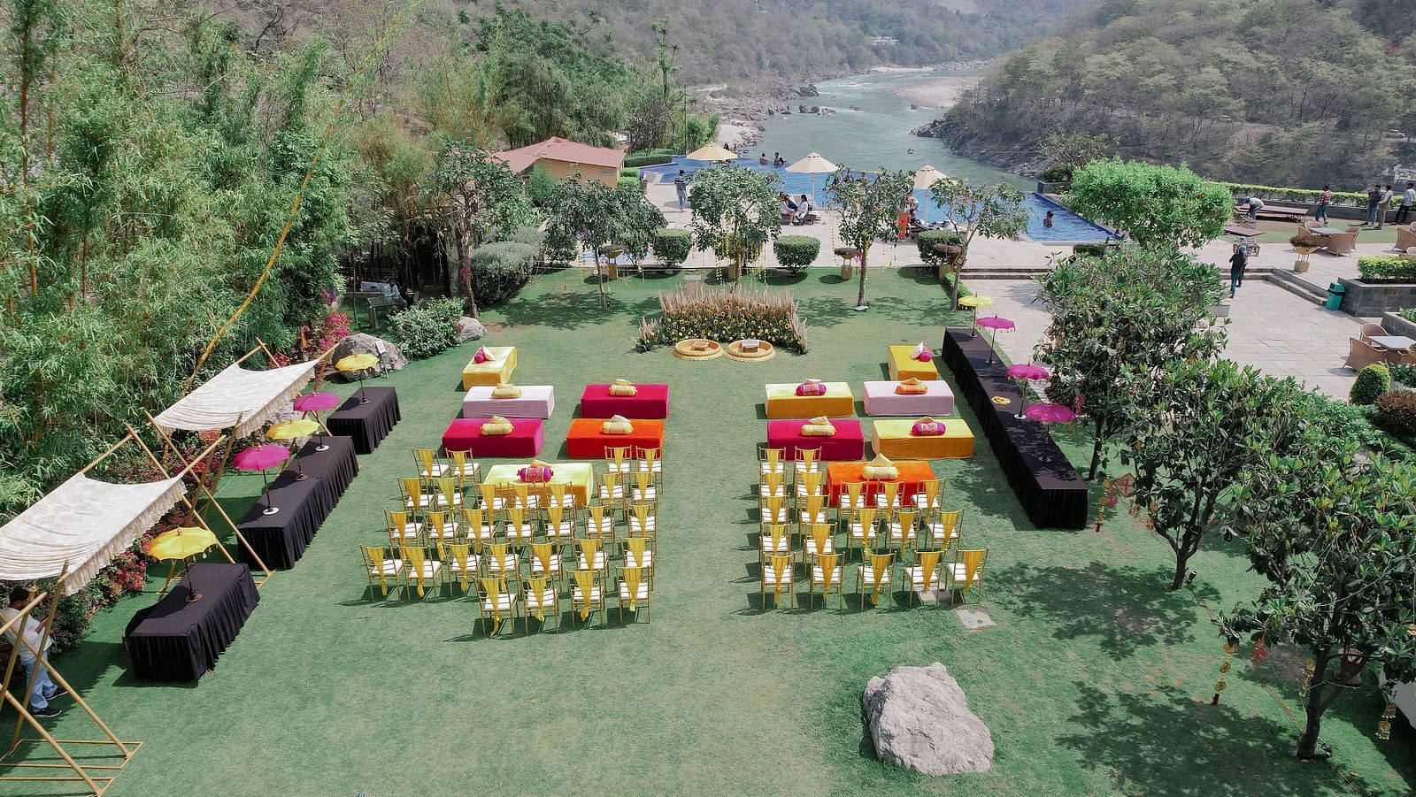 Aerial view of the outdoor venue. The lawn is decorated with colorful seating arrangements, including rows of chairs and square seating platforms. A large floral decor setup is visible in the center