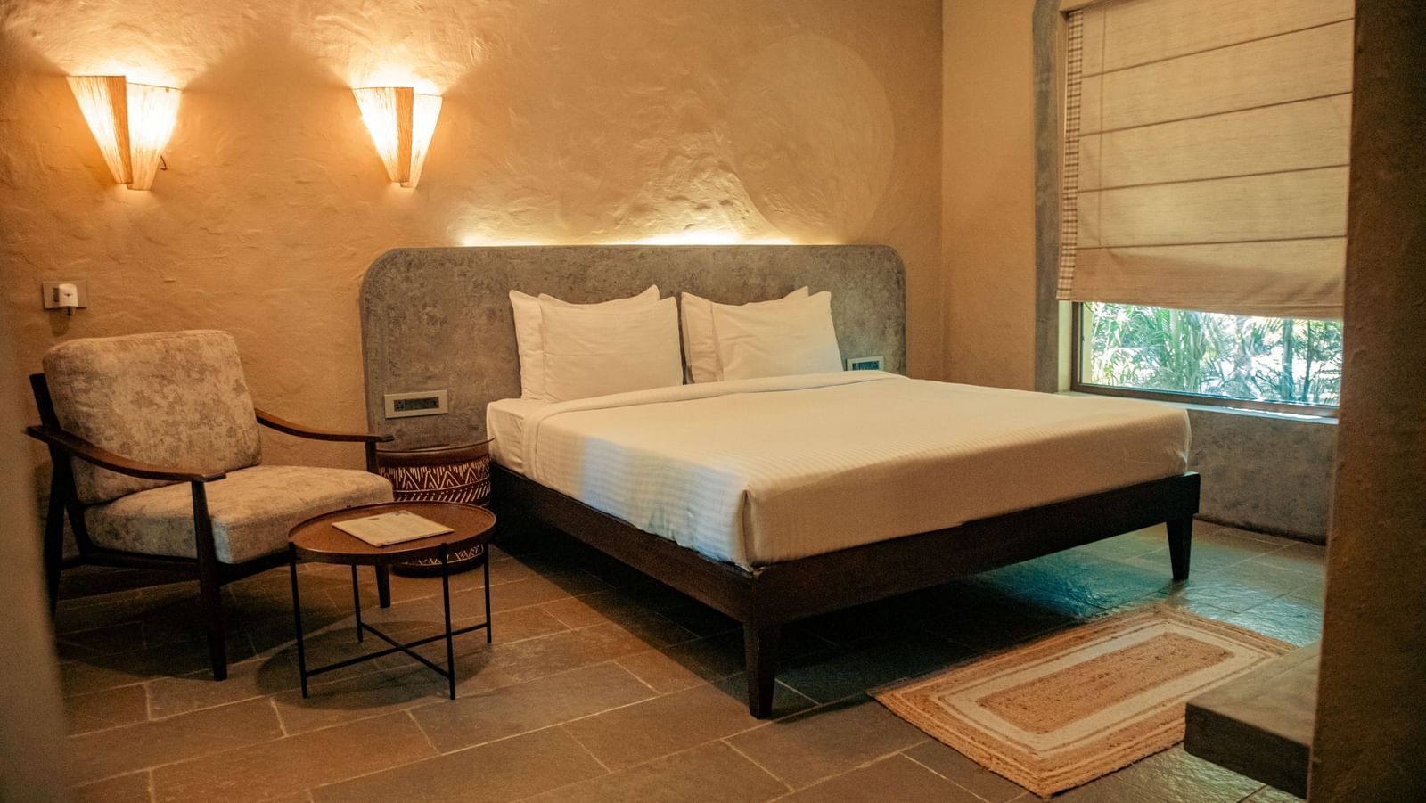 Cosy bedroom with a king size bed, an armchair, side table and wall lights against a soft beige wall at Fireflies Resorts, Kabini.