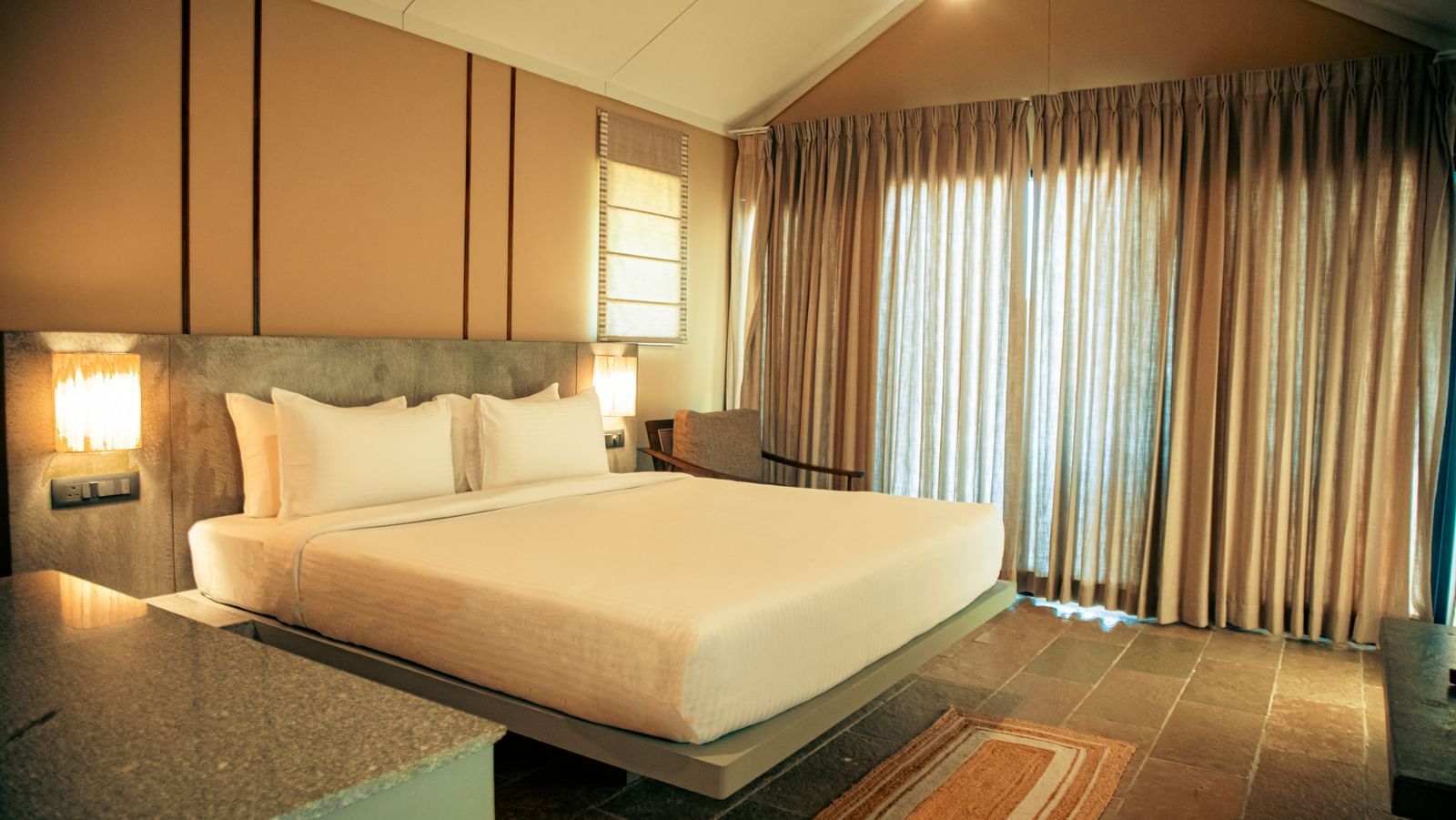 Bedroom with king size bed, bedside lamps, cushioned headboard, and full-length curtain covering a large window at Fireflies Resorts, Kabini.