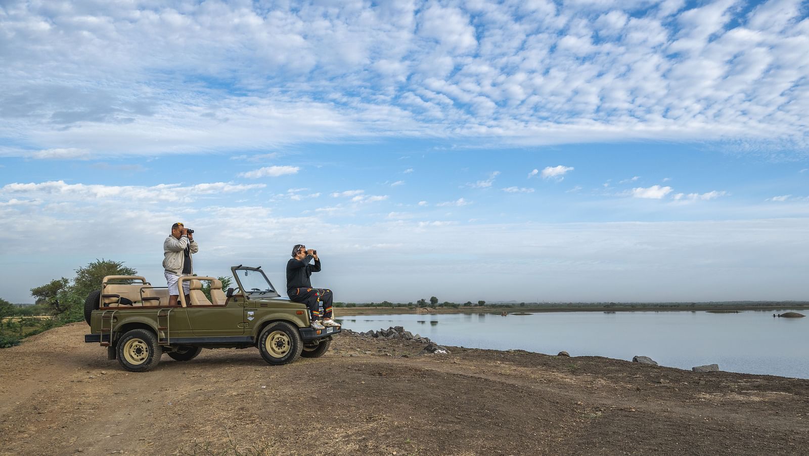 People stand in a vehicle, observing a waterside landscape under a partly cloudy sky - image provided by Fort Barli, Ajmer.