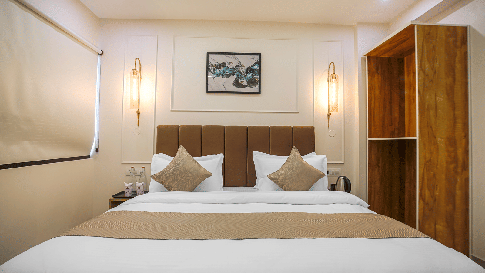 A neatly made king size bed at Perfectstayz Premium Varanasi, featuring a cushioned headboard, an artistic wall frame, and a wooden carved cupboard beside the bed.