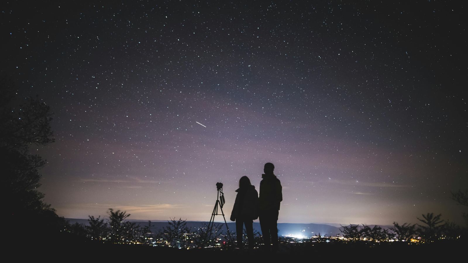 A wide view of two people standing beside a telescope under a starry sky.