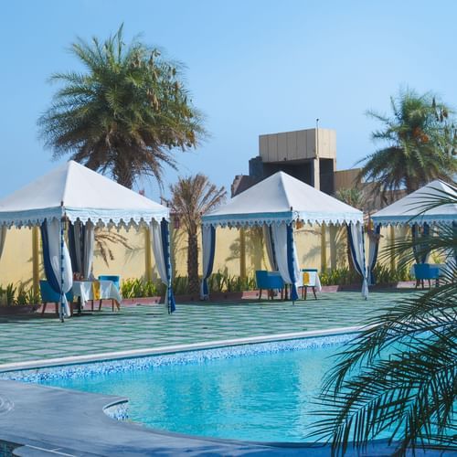 A serene poolside setting with white cabanas and palm trees, offering a relaxing environment under a clear blue sky - The Garden Ananta Elite, Rajkot