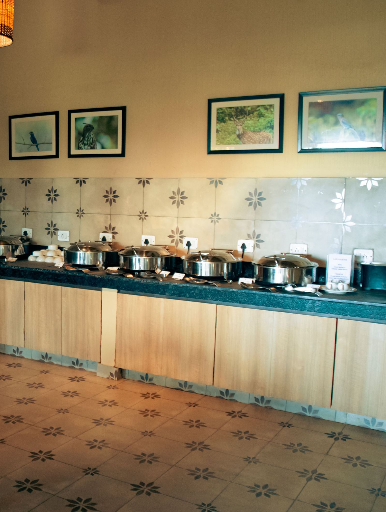 Breakfast buffet with chafing dishes and dispensers lined along a corner counter in a tiled room at Fireflies Resorts, Kabini.