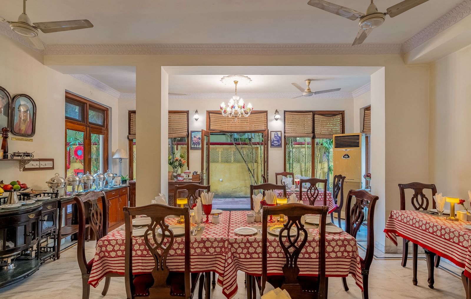The spacious dining hall at Dera Rawatsar, Jaipur, offers set tables with patterned cloths, a buffet setup, and a view towards an outdoor area.