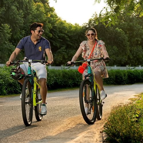 Two guests riding bicycles along a green path with trees on either side at Heritage Village Resorts & Spa, Manesar.