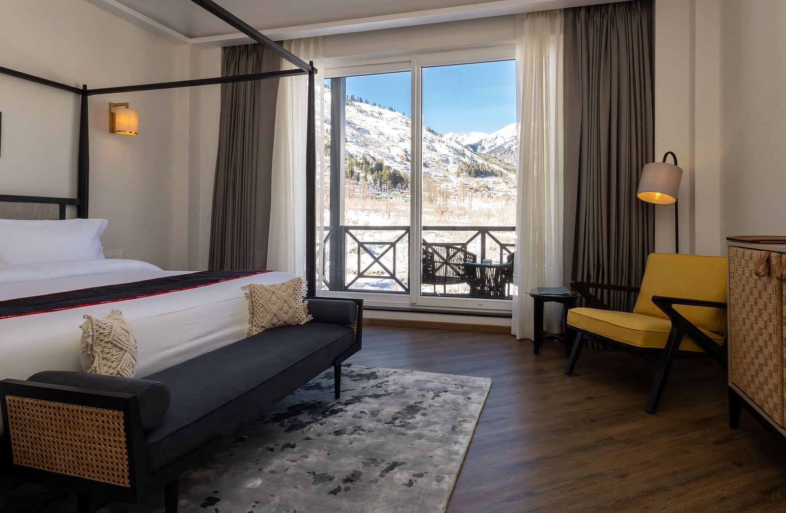 Diagonal view of the bedroom with a spacious bed, wooden seating at the foot, and large glass door opening to serene nature views from the balcony at Nivasana Manali.