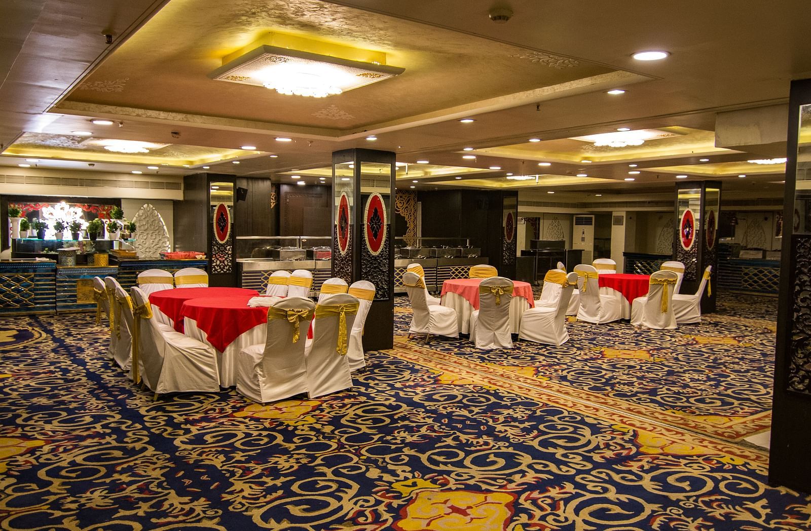 Maurya featuring round tables draped in red cloths and surrounded by white chairs, featuring a patterned carpet and decorative pillars.