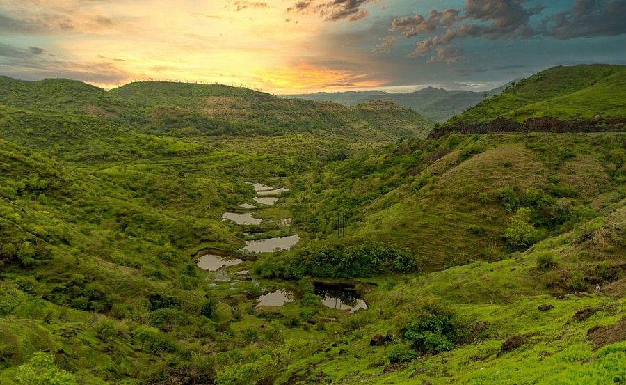 A panoramic view of a lush green valley dotted with water bodies at sunset, showcasing a dramatic sky painted with warm hues.