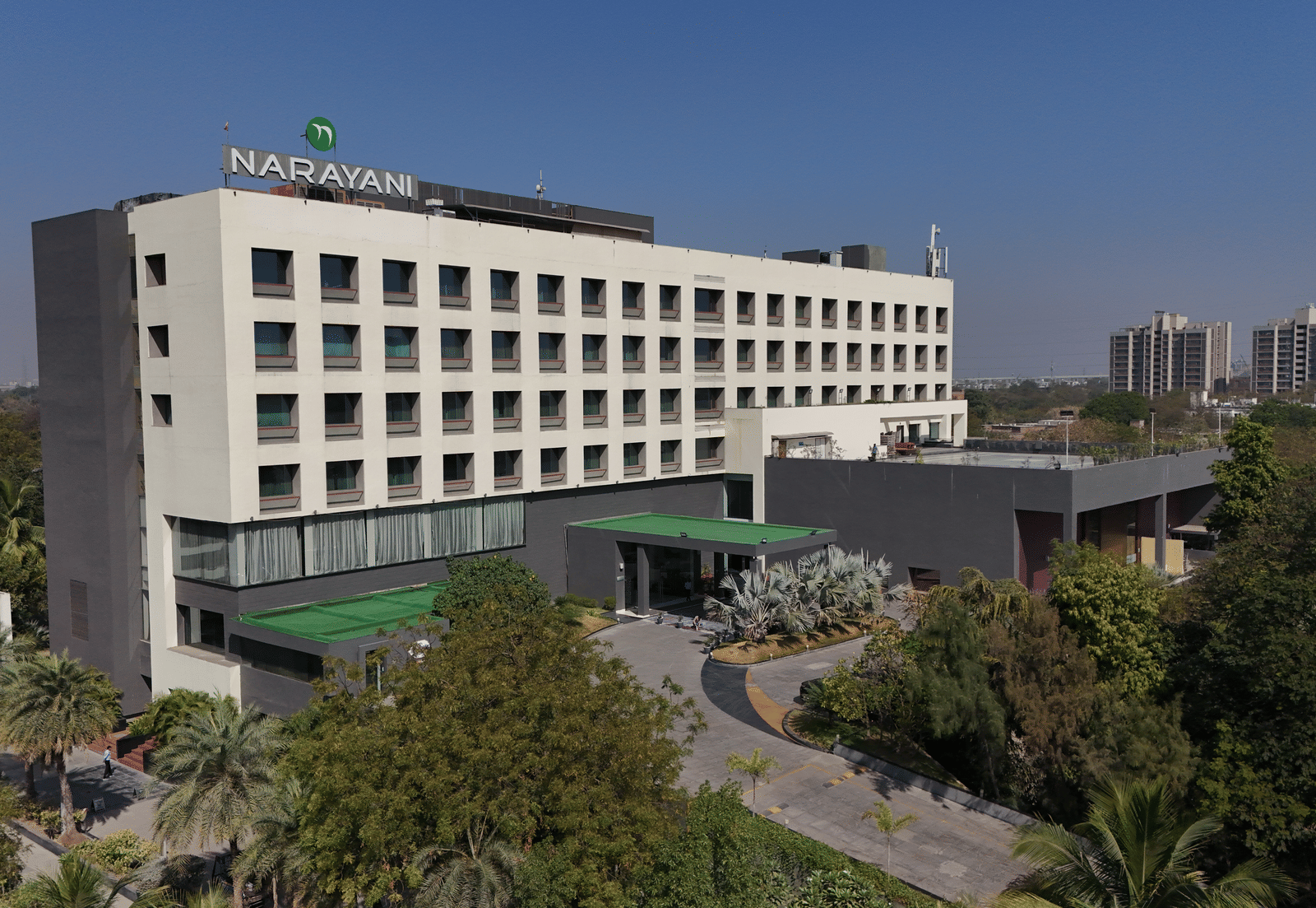 Long-shot of Narayani Heights Hotel & Resorts, Ahmedabad facade with trees surrounding it under a clear sky.