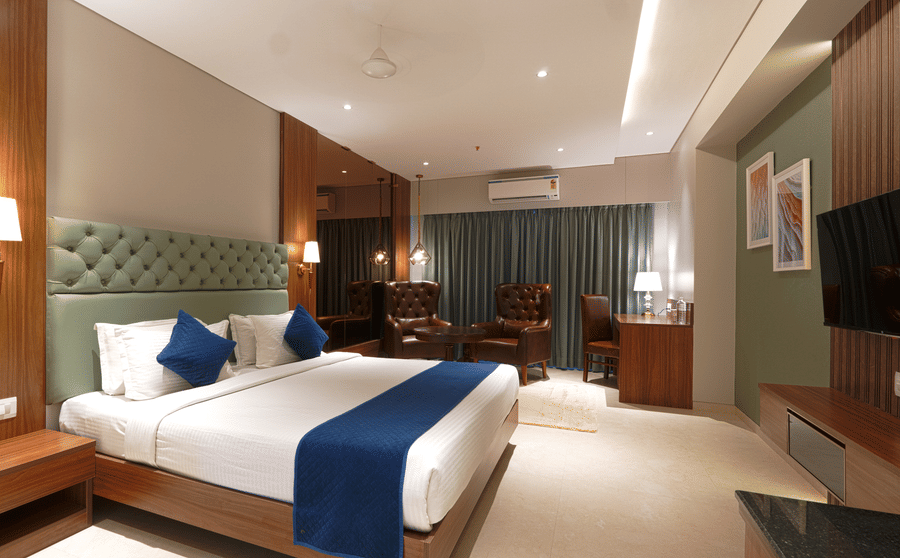 A room featuring a king size bed with a green headboard, a wooden desk, a television, and a brown leather armchair by the window at Magnus Kapitol, Sakore Nagar, Viman Nagar.