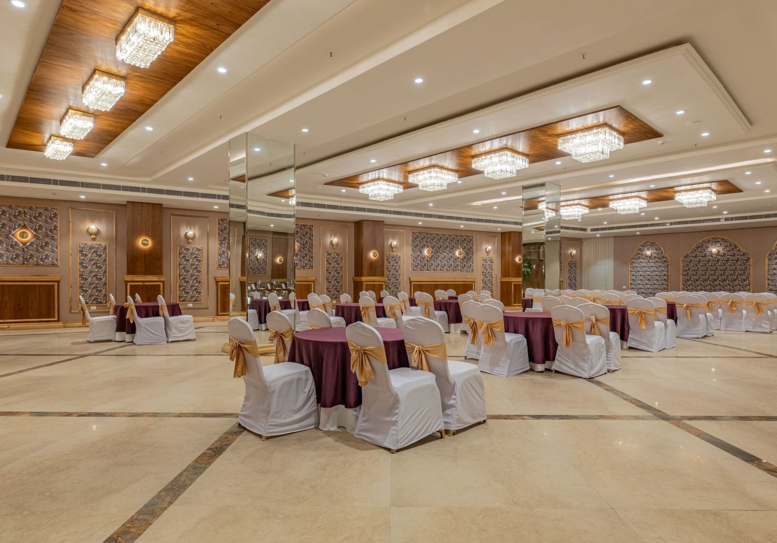 A large, elegant banquet hall is arranged with numerous round tables draped in white and purple, featuring a central aisle and decorative pillars.