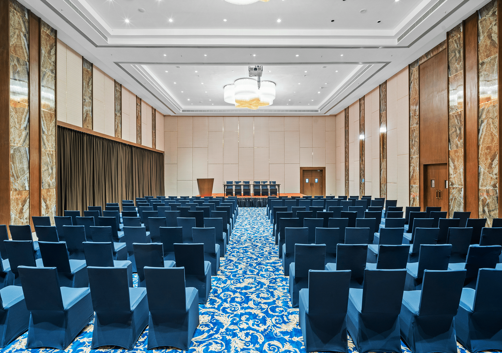 An image of Champavati Banquet Hall with blue chairs facing towards a podium at Grand Vijayawada by GRT Hotels.
