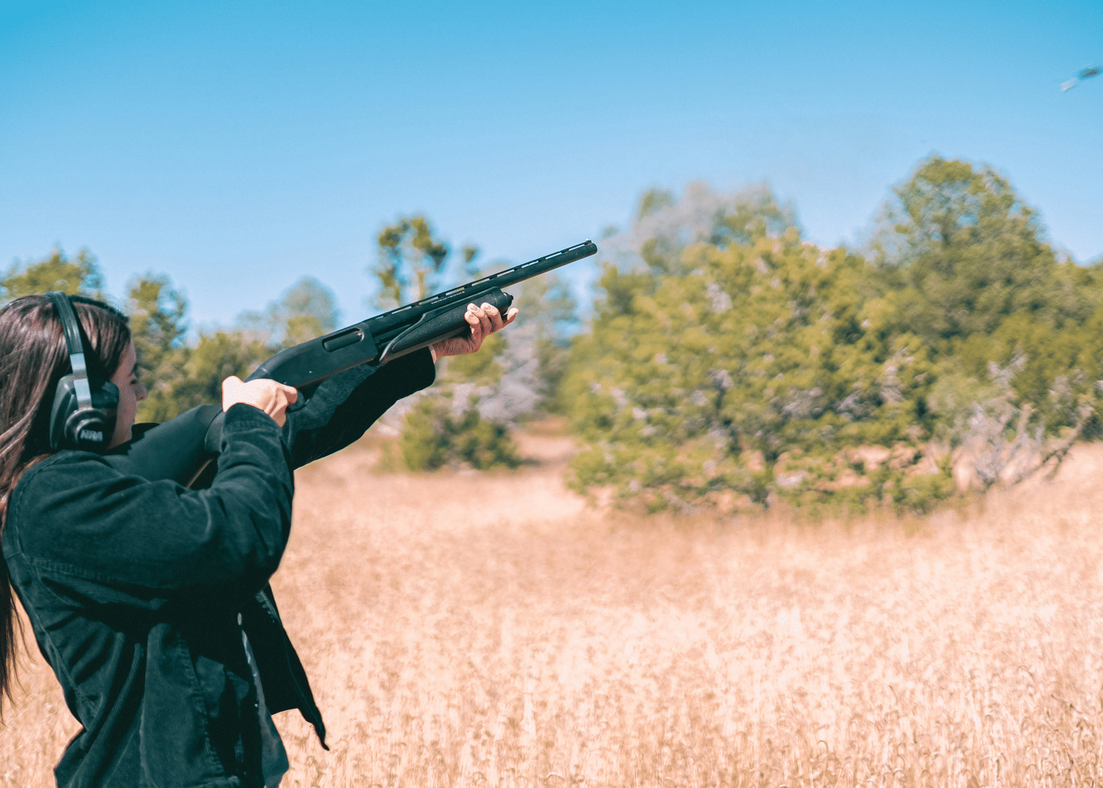 A person, seen from behind, aiming a rifle across a dry, reedy landscape under a clear, blue sky.