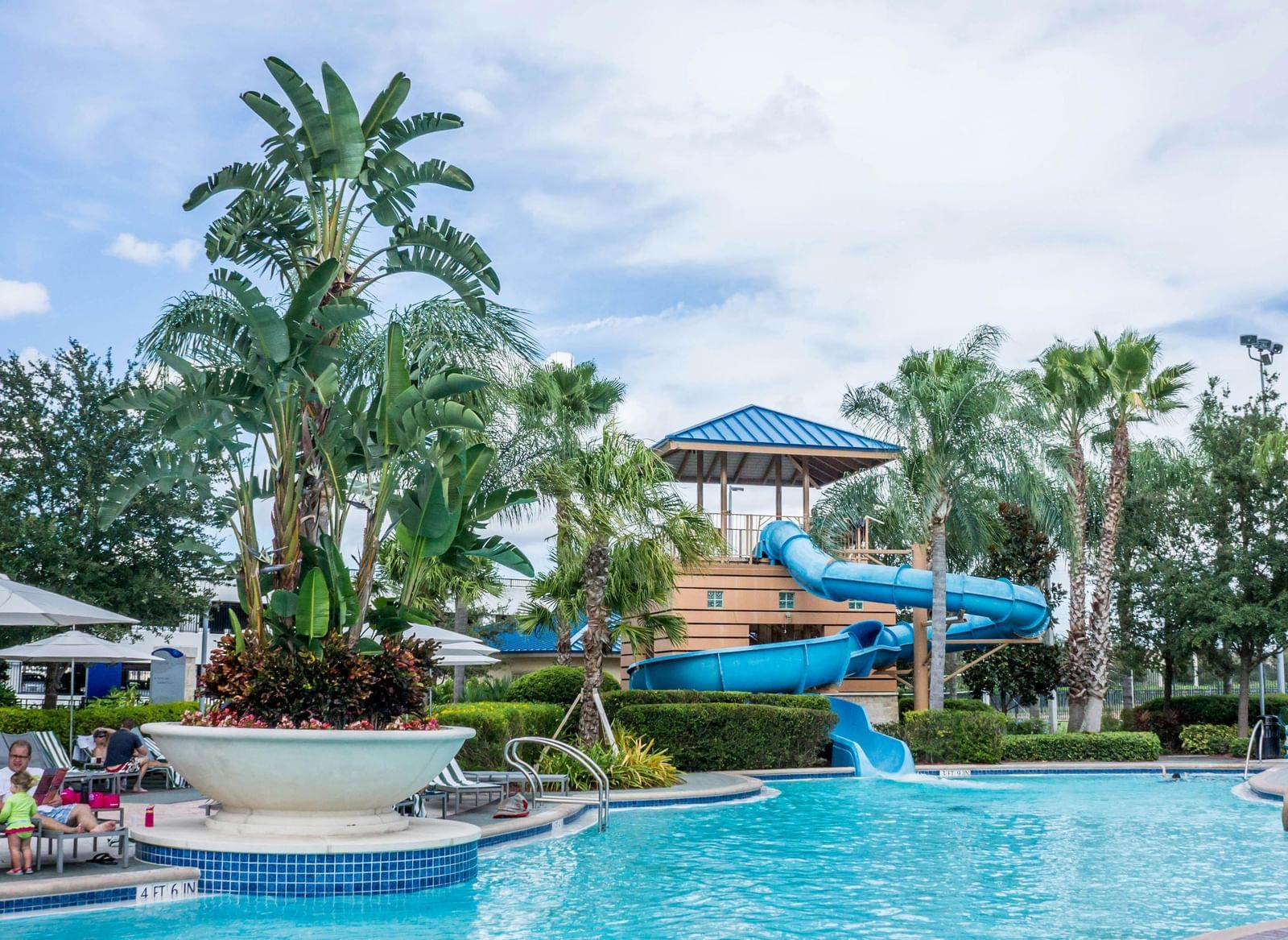 A swimming pool with palm trees, lounge chairs, and a blue water slide in the background.