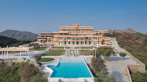 Palace-style hotel exterior with a central swimming pool, landscaped gardens, stone pathways, pavilion structures, and surrounding hills under a clear blue sky - Fateh Vilas, Udaipur
