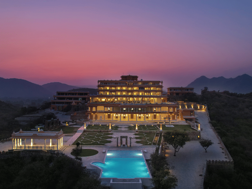Aerial view of Fateh Vilas, a heritage resort in Udaipur, during twilight hour, with a swimming pool, a large lawn area, a canopy, walking pathways, and mountain in the background seen
