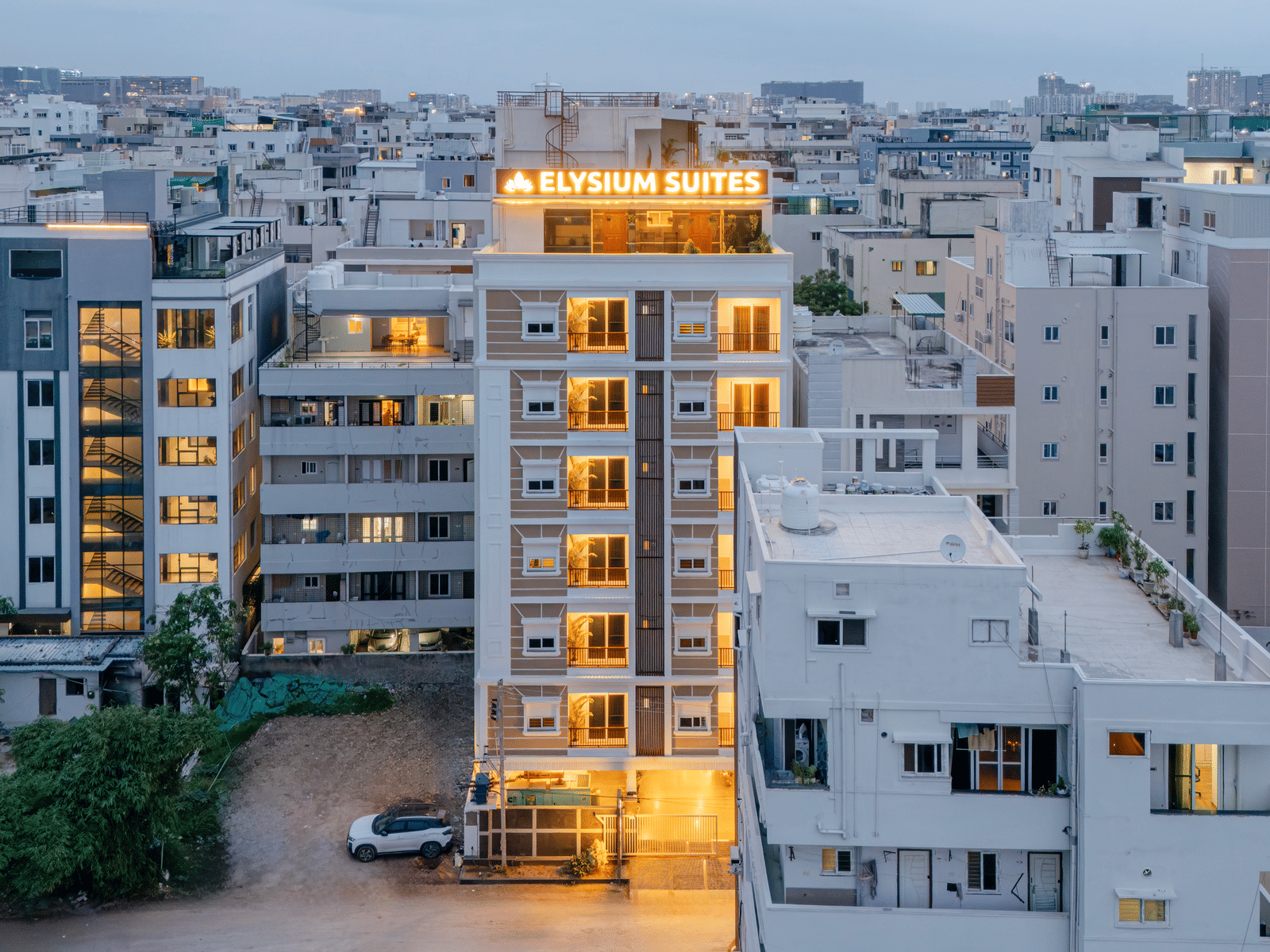 Buildings stand close together in a city at dusk with lights visible from windows as night falls near Elysium Studio Suites, Madhapur..