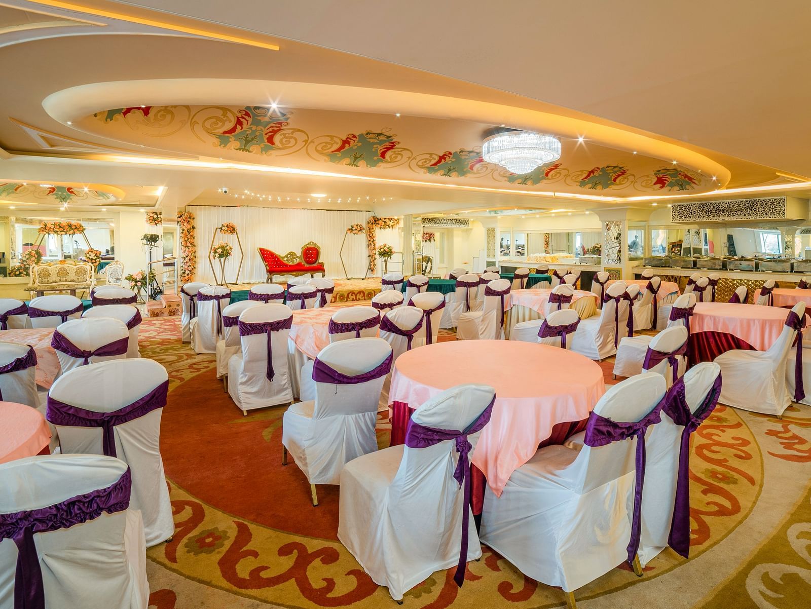 Samrat featuring round tables draped in pink tablecloths and chairs adorned with white covers and purple sashes.