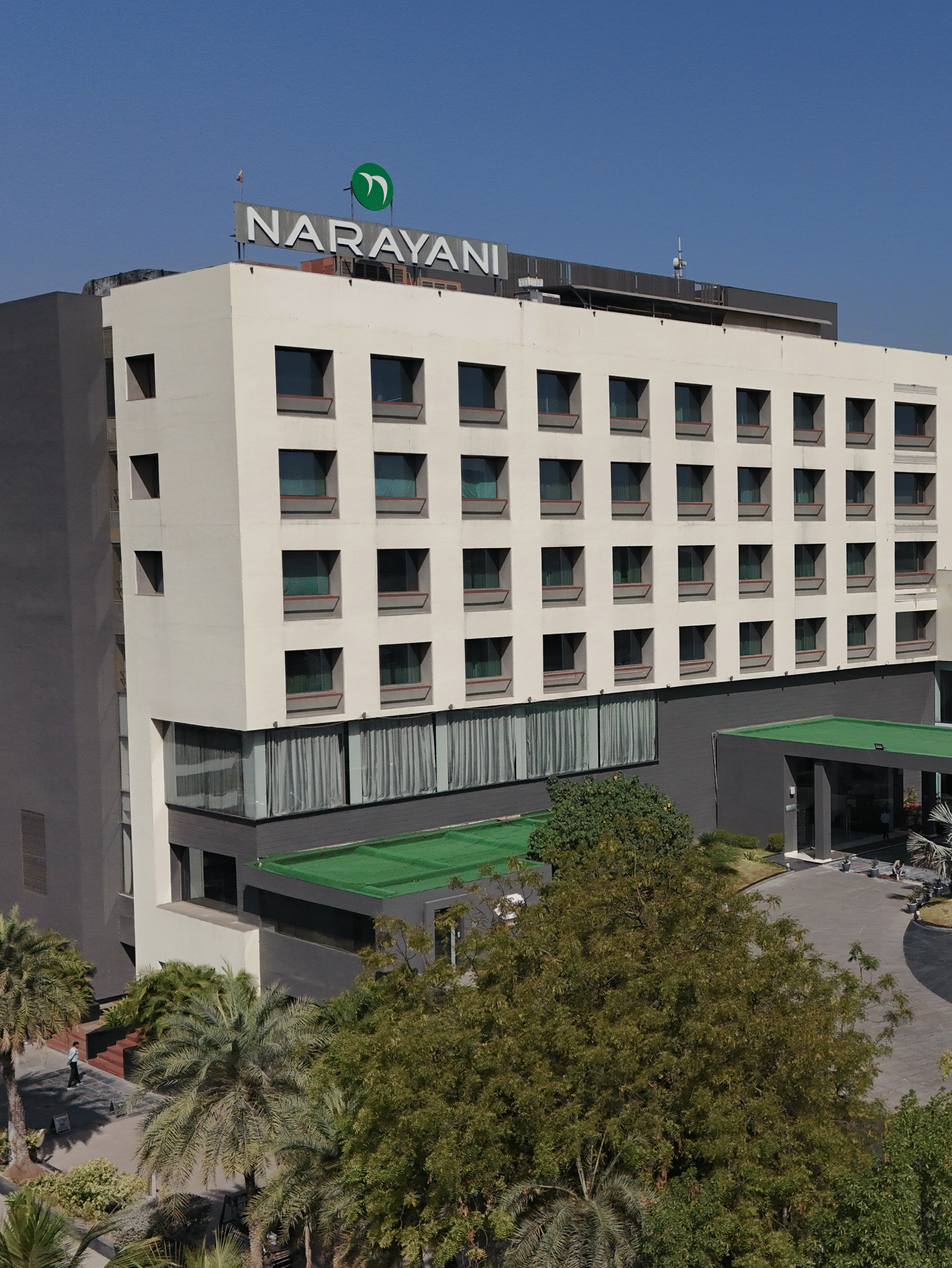 Long-shot of Narayani Heights Hotel & Resorts, Ahmedabad facade with trees surrounding it under a clear sky.