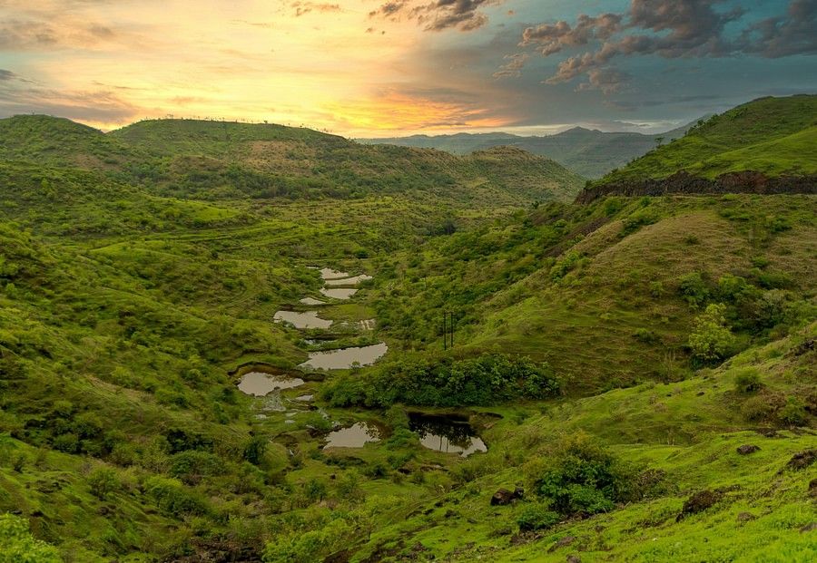 alt-text A panoramic view of a lush green valley dotted with water bodies at sunset, showcasing a dramatic sky painted with warm hues.