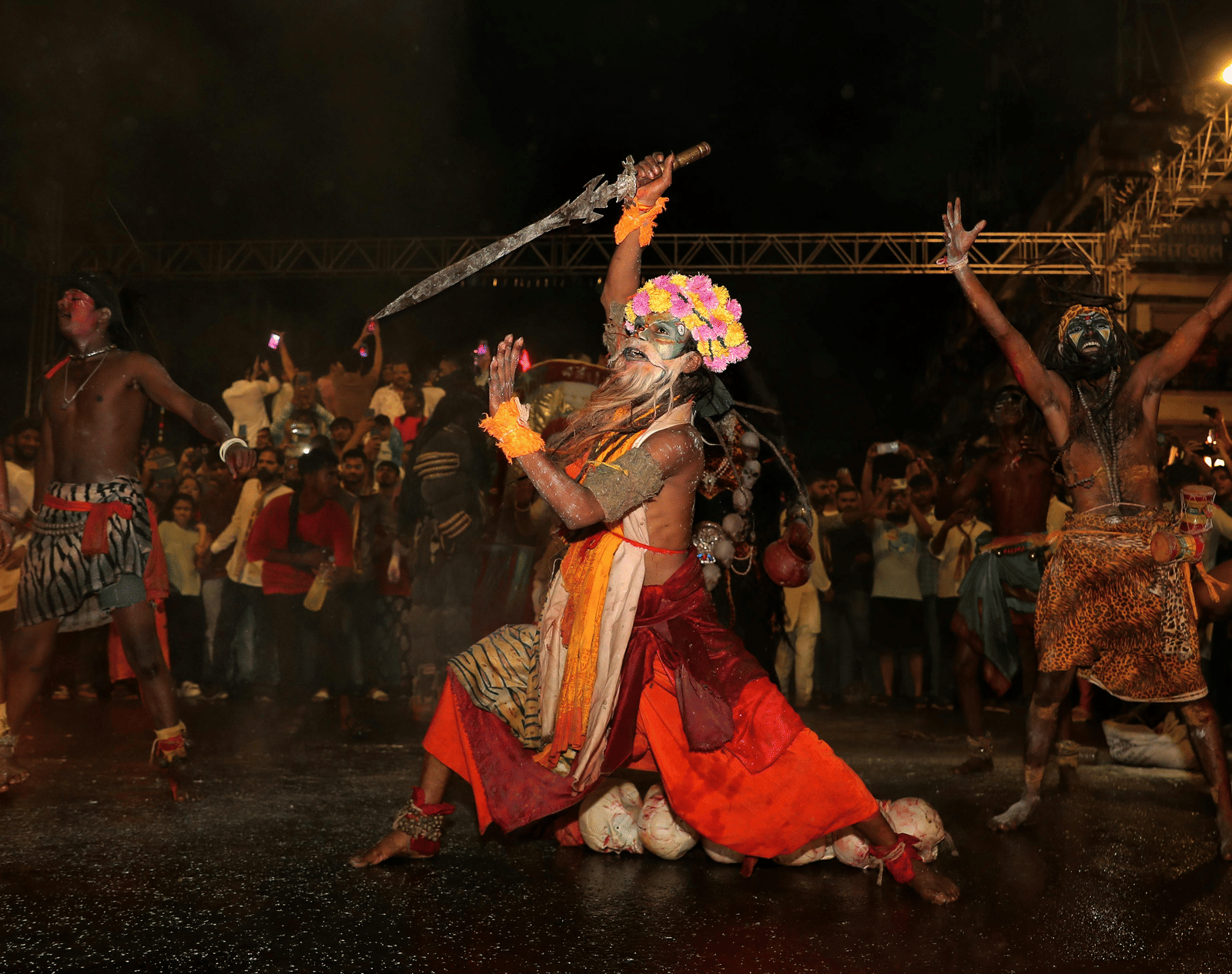 Traditional cultural folk dance performance taking place at night with performers in colourful costumes, face paint and dramatic poses during a festive celebration.