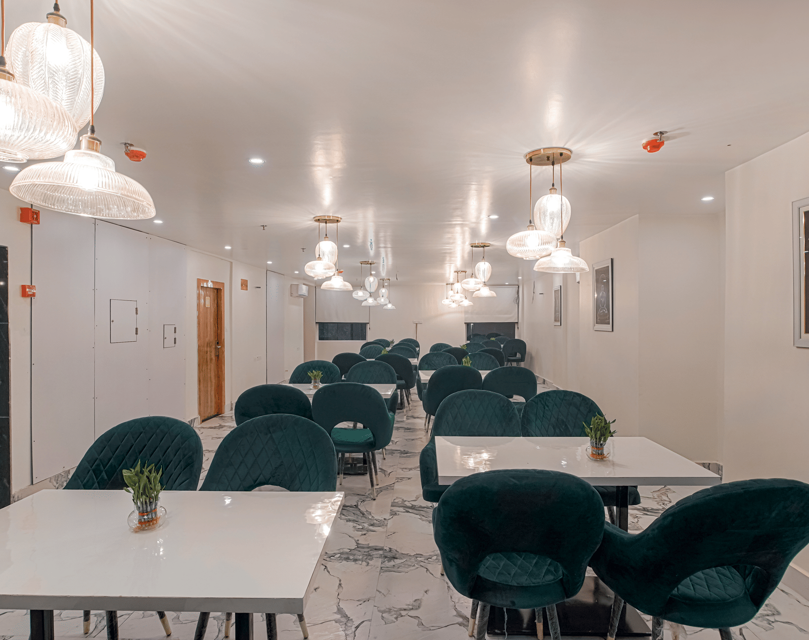 A clean, well-lit view of the modern dining room at Perfectstayz Premium Varanasi, featuring dark seating, white tables, an artistic wall frame, and marble flooring.