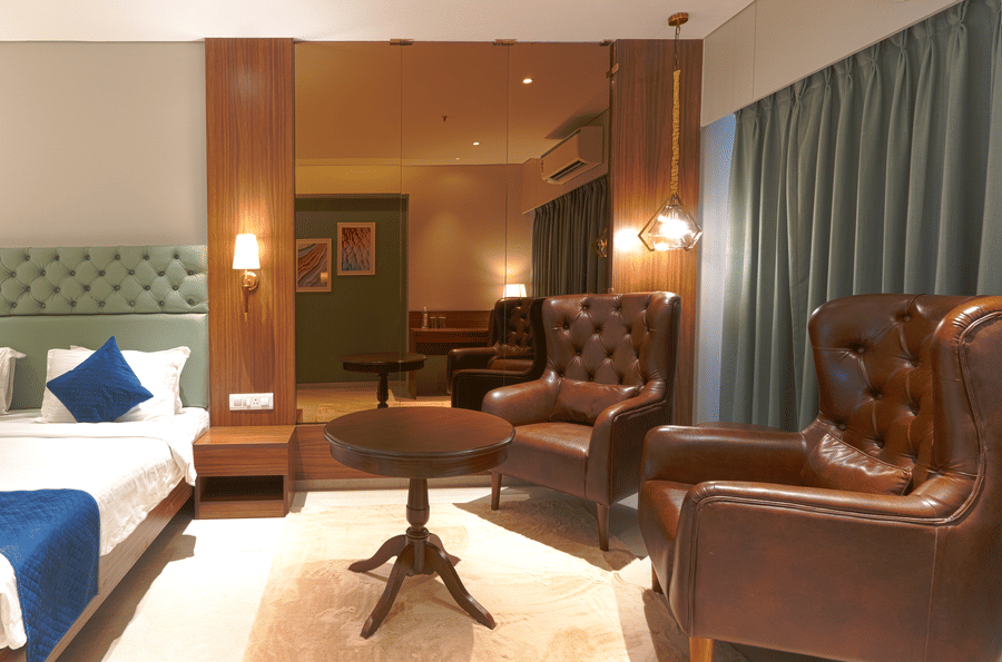 A room with a bed featuring green headboard, white bedding with a blue runner, a small round table, and a brown leather armchair at Magnus Kapitol, Sakore Nagar, Viman Nagar.