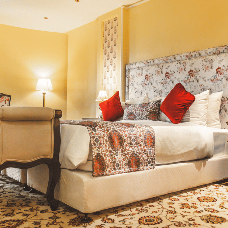 The energised look of the Super deluxe cottage at The Garden Ananta Elite with enchanting white table lamps on both sides, red pillows to up the comfort, and a cushioned seat for two, placed adjacent to the bed.