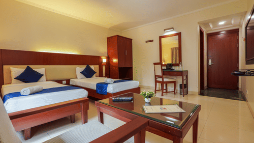 A neat hotel room with two single beds, blue accents, a wooden desk, a wardrobe, a small seating area with a glass table, and a door leading to the hallway - Magnus Riverside, Kalyani Nagar