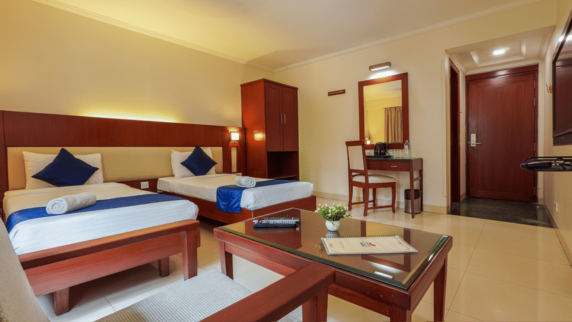 A neat hotel room with two single beds, blue accents, a wooden desk, a wardrobe, a small seating area with a glass table, and a door leading to the hallway - Magnus Riverside, Kalyani Nagar