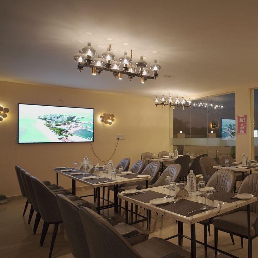 A stylish restaurant with elegant grey chairs, marble tables, and soft lighting. Large windows offer a view of the outside greenery at Citrus Classic Resorts and Spa Mahabalipuram ECR