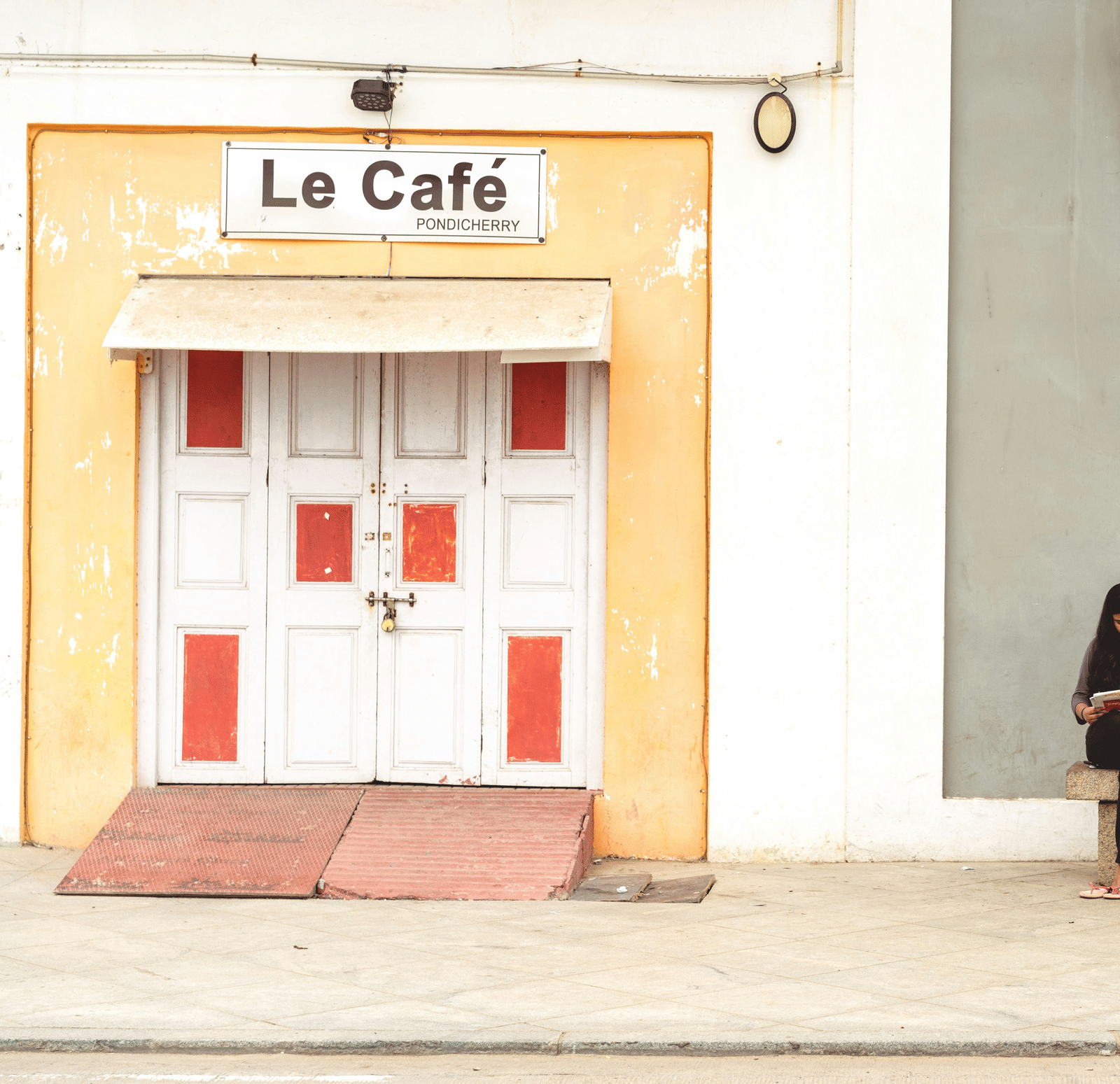 A person sits on the steps of a yellow building with a sign that reads 'Le Cafe' and a blue shuttered window.