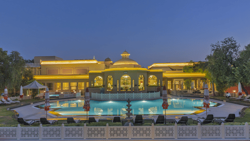 The facade of Heritage Village Resorts & Spa, Manesar, featuring the brightly illuminated exterior along with an expansive swimming pool in the foreground under the dark blue skies, perfect for a summer staycation near Delhi.