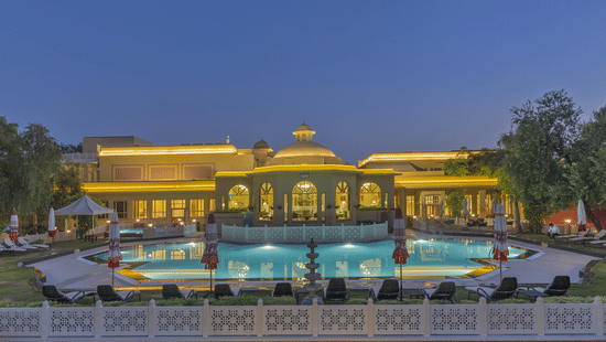 The facade of Heritage Village Resorts & Spa, Manesar, featuring the brightly illuminated exterior along with an expansive swimming pool in the foreground under the dark blue skies, perfect for a summer staycation near Delhi.