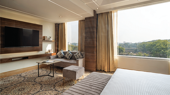 A spacious room at Clarks Inn Suites, Near Taj Mahal featuring a large TV mounted on the wall next to which a sitting space is arranged with sofa and a pouffe around a coffee table near the large window