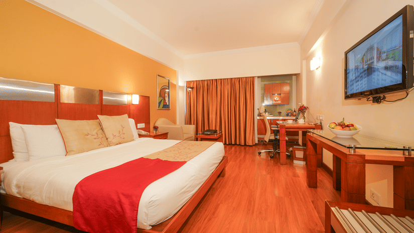 Luxury suite bedroom at The Suryaa New Delhi featuring a large bed with white linens, a beige runner, a red blanket, side tables, and wall-mounted lamps and with curtains hanging