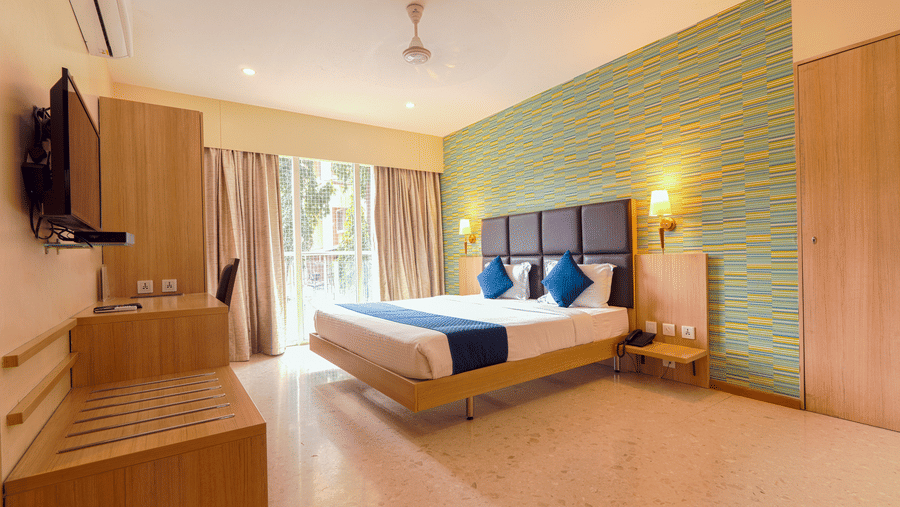 A well-lit, modern hotel room at Magnus Star Residency, Koregaon Park Annexe, featuring a double bed with blue accents, a stylish patterned wall, work desk, mounted TV, and a large window with curtains.