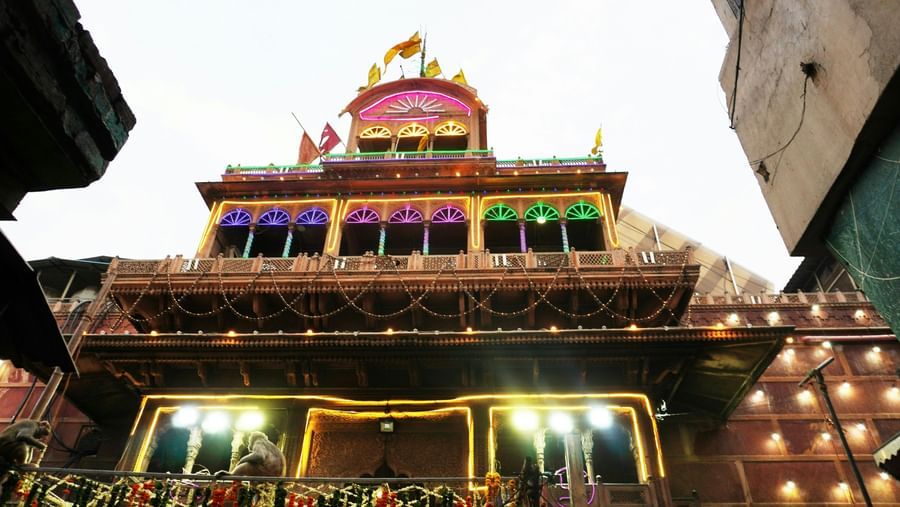 Beautifully lit temple facade at dusk with intricate carvings, arched windows, colourful lights, hanging garlands, and monkeys sitting near the entrance.