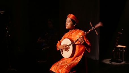 A musician in traditional attire playing a sitar on a stage, with a dark background.