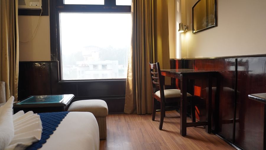 A bedroom with a chair and a table, bathed in light streaming through the window at DLS Park Grace, Haridwar.