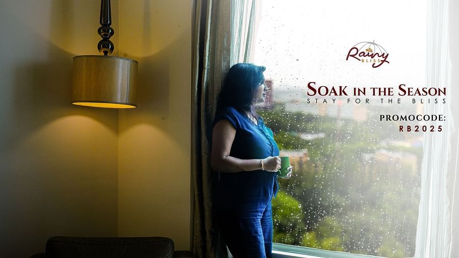 A woman enjoying a warm beverage while gazing out of a hotel window on a rainy day, evoking comfort and tranquility. Promotional offer text on the image reads, Rainy Bliss – Soak in the Season. Stay for the Bliss. Promocode: RB2025