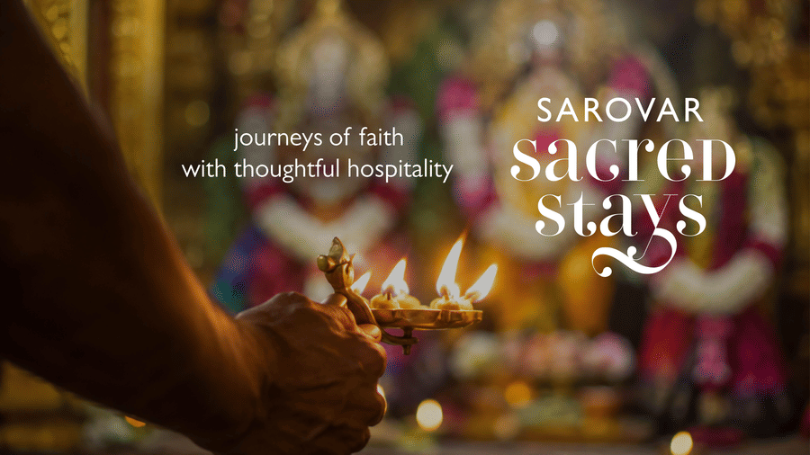 A hand holds a lit oil lamp in a temple, with blurred deities in the background with caption Journeys of faith with thoughtful hospitality, Sarovar Sacred Stays.